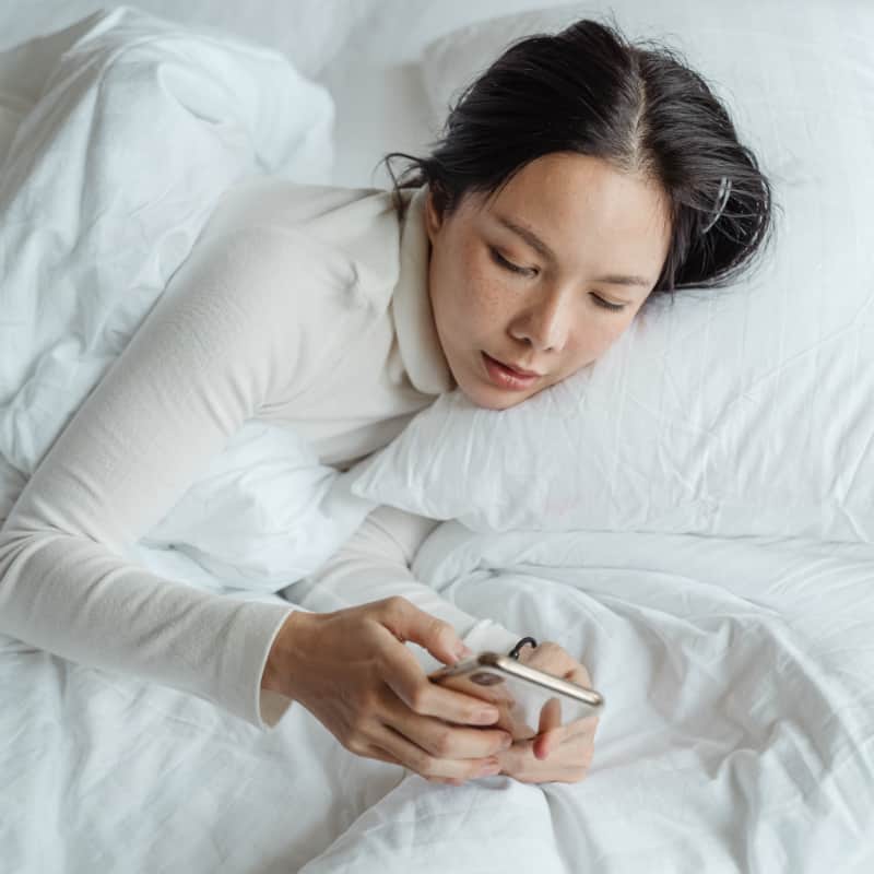 A woman reading texts on her phone while lying in bed, representing the need to establish boundaries around technology use and self-care.