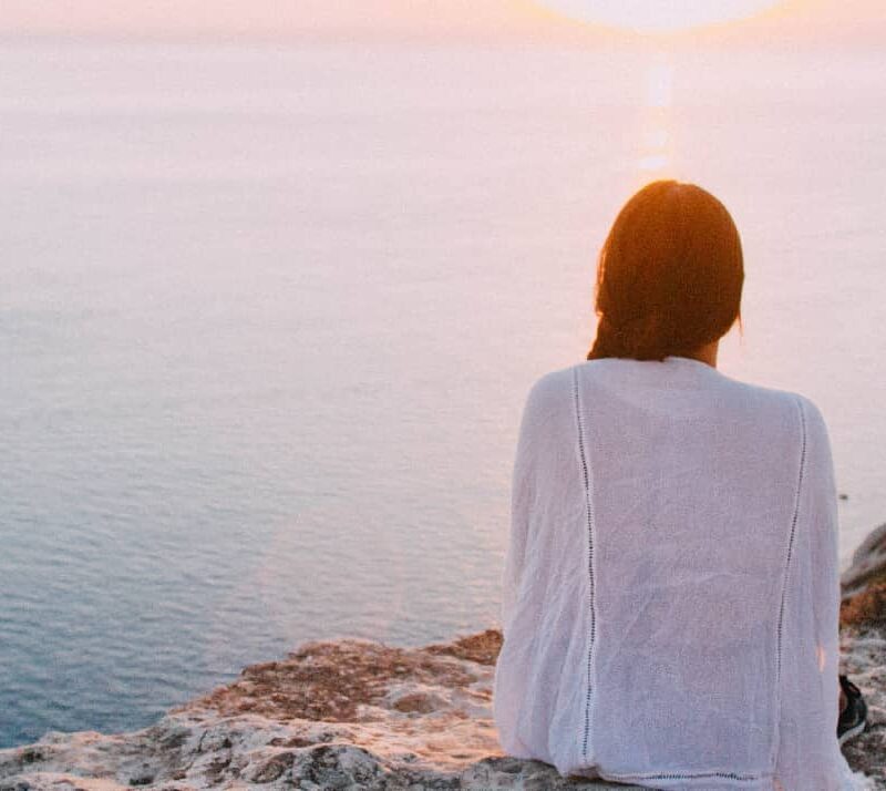 A woman standing on a hill, gazing thoughtfully at a serene sea during sunset symbolizing the journey of long term mental health recovery and the calmness that comes with long-term care.